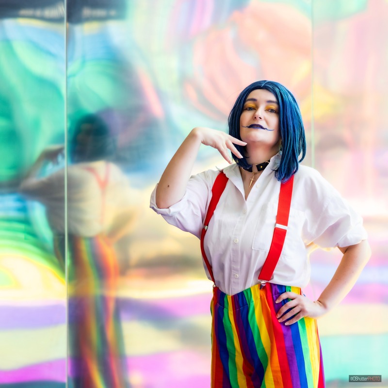 MDA dressed in humanized Asmodeus. Blue wig, blue lips extended out. White dress shirt with red suspenders and rainbow pants. Reflective colourful wall in behind. They're standing with one hand on their hip and the other by their chin.