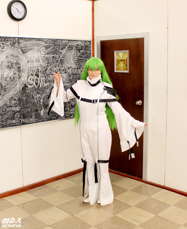 MDA dressed as CC from the anime Code Geass in her white bodysuit with black straps. Standing towards the corner of a fake room with the door to the right and a blackboard covered in chalk to the left
