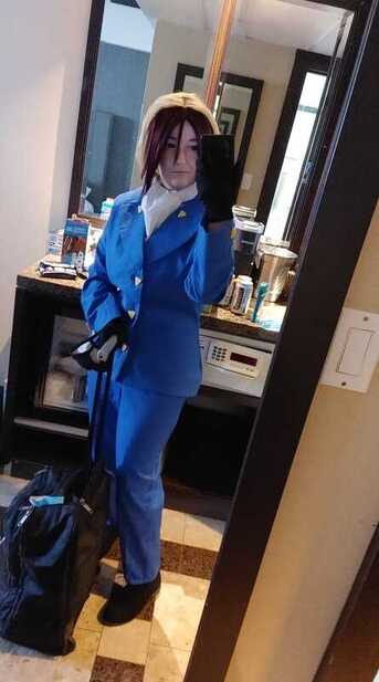 MDA dressed as Nougami Neuro from the Majin Tantei Nougami Neuro. Mirror selfie and they're looking at their phone. A bright blue two piece suit with yellow triangle buttons down the front of the jacket, a white cravat around their neck. They hold their backpack handle in their right hand.